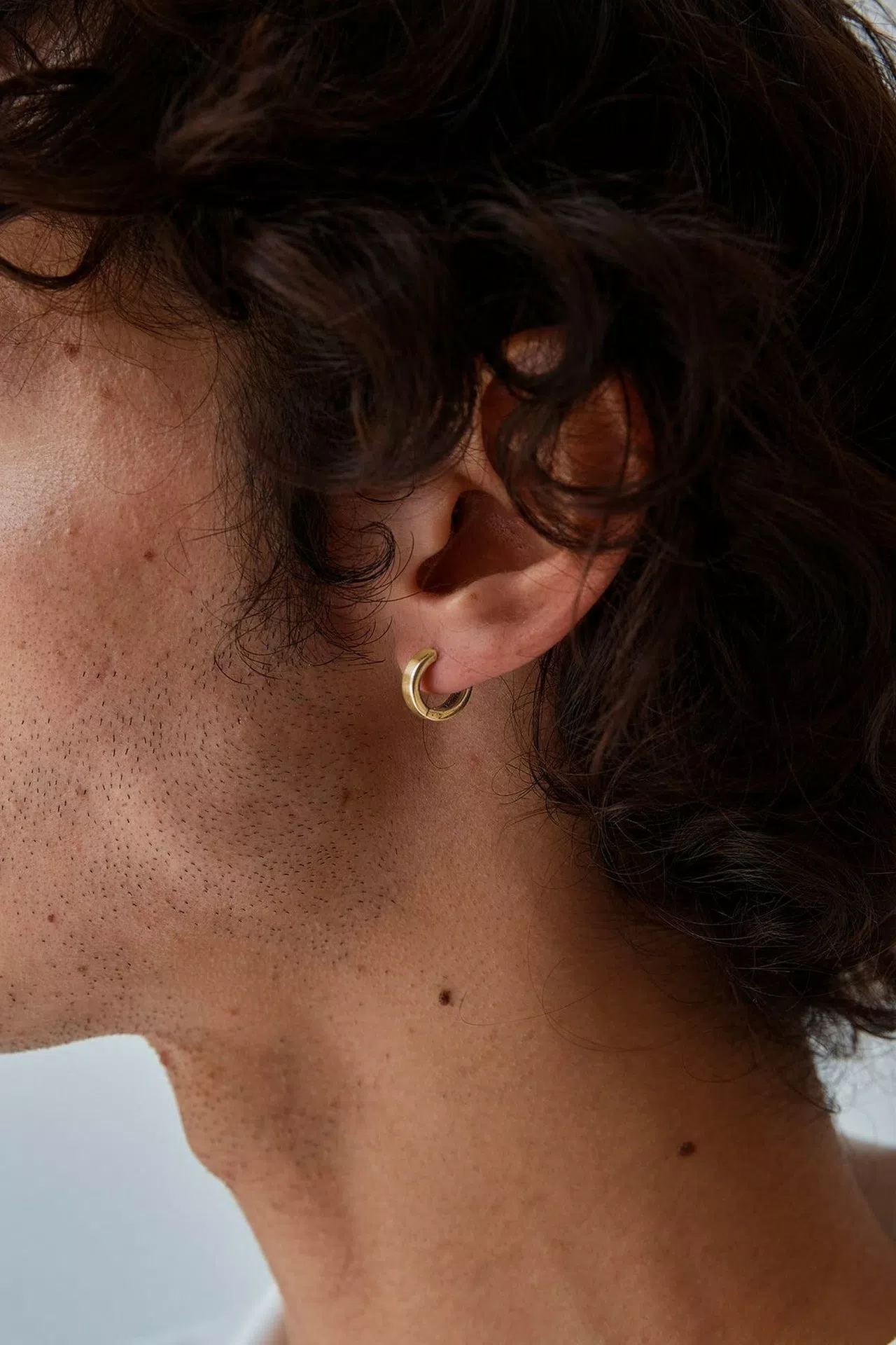Gros plan d'un lobe d'oreille d'un homme portant une petite créole dorée et simple. Ses cheveux bruns bouclés sont visibles sur l'arrière-plan.